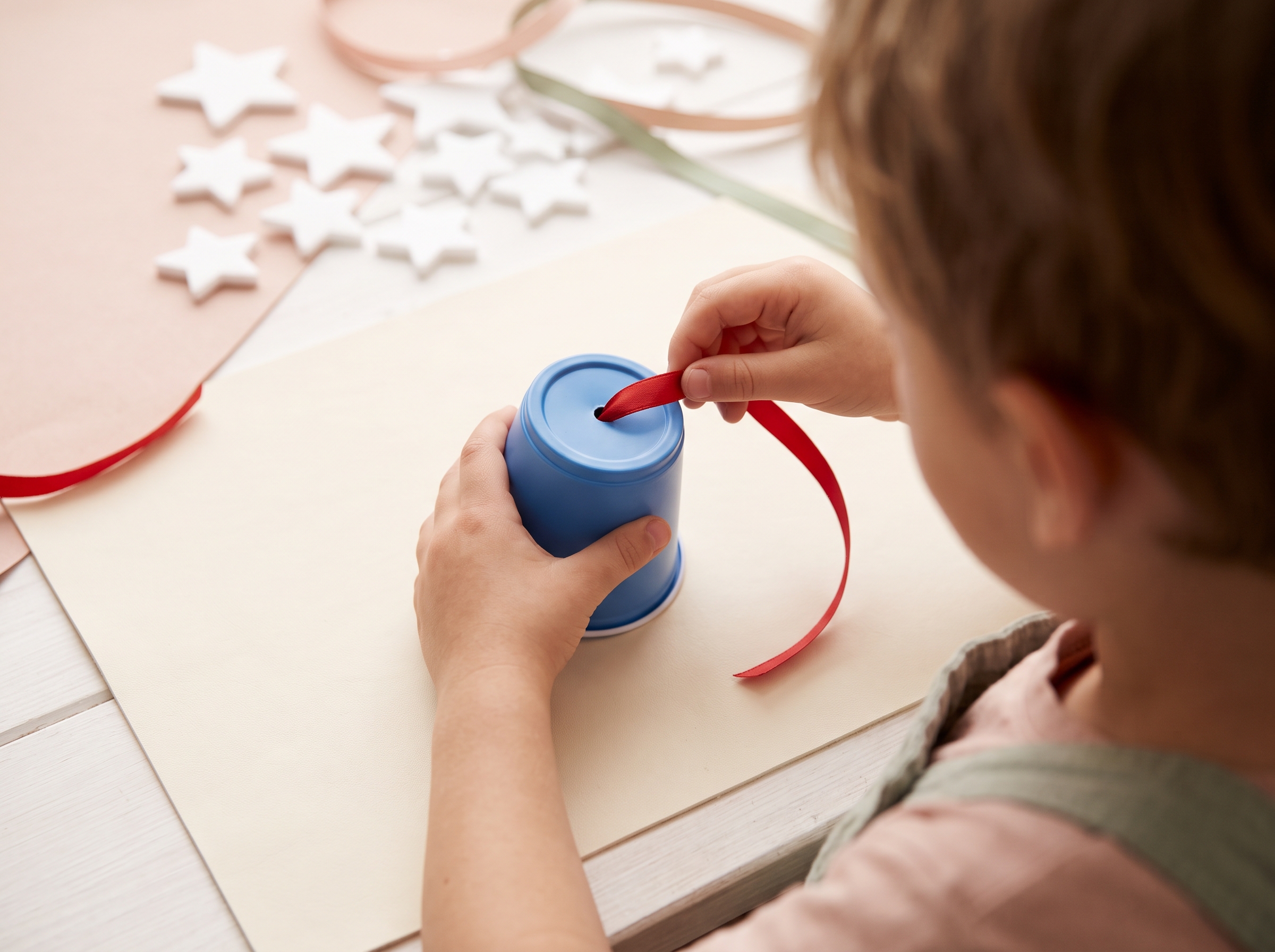 Child's hands threading a red satin ribbon strip through a hole punched in a blue plastic cup