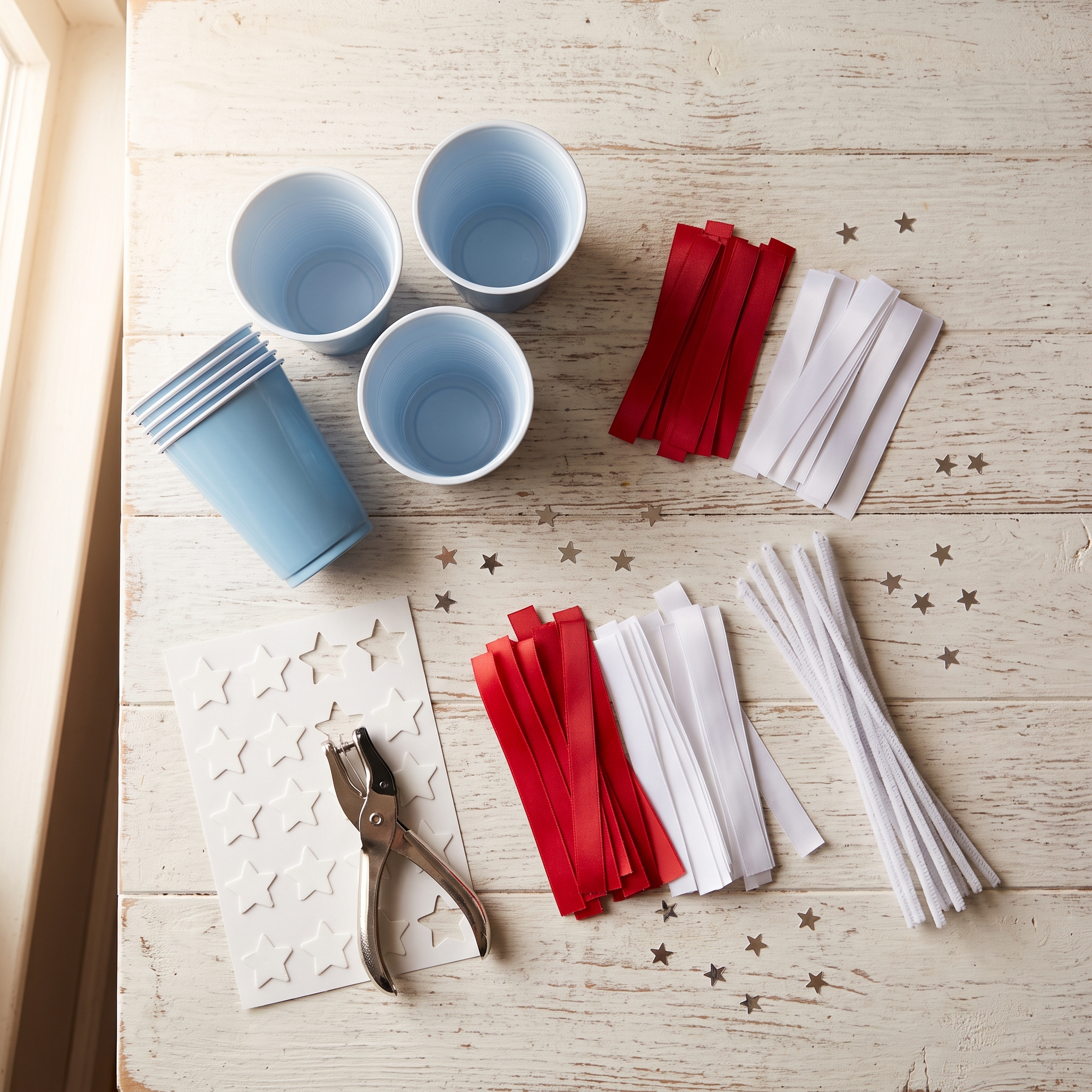 Craft supplies arranged on a white surface: blue plastic cups, red and white satin ribbons, white foam star stickers