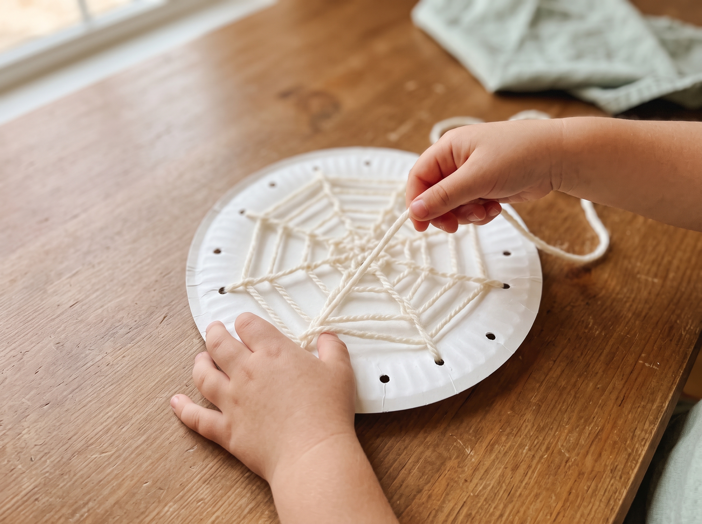 Child's hands threading cream yarn through holes punched in a white paper plate to create a spider web pattern